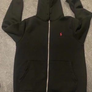 Svart hoodie från Polo Ralph Lauren - Svart hoodie från Polo Ralph Lauren med dragkedja och huva. Tröjan har en röd broderad logga på bröstet och två stora fickor framtill. Tillverkad i 100% bomull för en skön och mjuk känsla. Perfekt för en chill och stilren look.