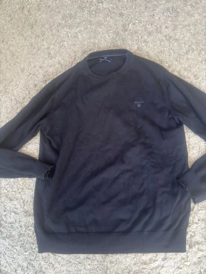 Svart sweatshirt från GANT - Klassisk svart sweatshirt från GANT med rund halsringning och diskret logga broderad på bröstet. Tröjan har ribbade muddar vid ärmslut och nederkant. Perfekt för en clean och enkel stil.L men sitter mera som M