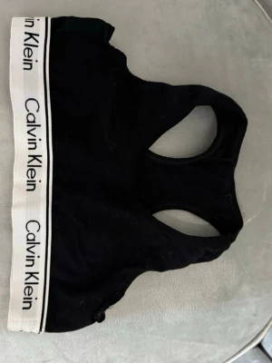 Svart sport-bh från Calvin Klein - Svart sport-bh från Calvin Klein med klassisk racerback och bred vit resår med logga runt om. Mjuk och bekväm modell i bomullsblandning, perfekt för en aktiv vardag eller som ett snyggt basplagg.