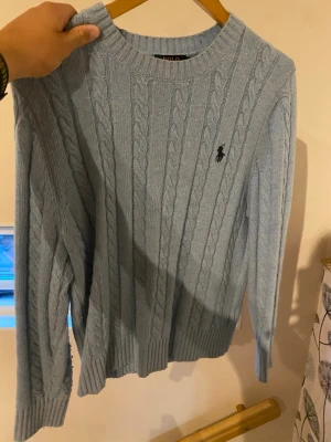 Ljusblå kabelstickad tröja Polo Ralph Lauren - Kabelstickad tröja i ljusblått från Polo Ralph Lauren. Klassisk rund halsringning, långa ärmar och ribbade muddar. Tröjan har den ikoniska Ralph lauren loggan på bröstet och är tillverkad i mjukt material som passar perfekt till kyligare dagar.