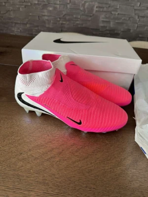Nike Phantom rosa elite fotbollsskor - Säljer ett par Nike Phantom fotbollsskor i en riktigt snygg rosa färg med vita och svarta detaljer. Skorna har strumpliknande skaft och är designade utan snörning för en tight passform. Sulan har fasta dobbar för spel på gräs. Perfekta för dig som vill sticka ut på planen! Helt nya och 100% äkta. Köpta ifrån intersport! Kvitto finns!!