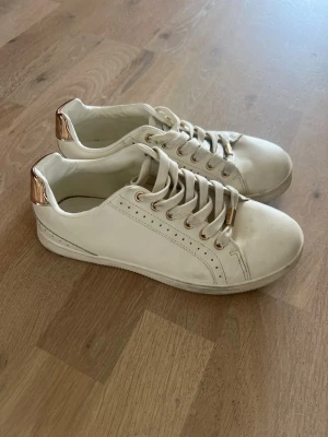 Vita sneakers med gulddetaljer, från ONLY - Snygga vita sneakers från ONLY,  med guldfärgade detaljer på hälen och snörhålen. Skorna har klassisk låg modell och är tillverkade i syntetmaterial med vita skosnören. Perfekta för en clean och stilren look.