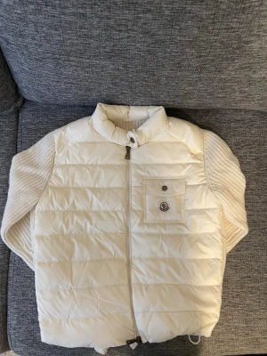 Vit cardigen  från Moncler - Snygg vit cardigen från Moncler med quiltad framsida, ribbstickade sidor och en bröstficka med knapp och Moncler-logga. Västen har hög krage, dragkedja framtill och justerbar nederkant med dragsko. Perfekt för lager på lager och streetstyle.Storleken är 1 vilket motsvarar S