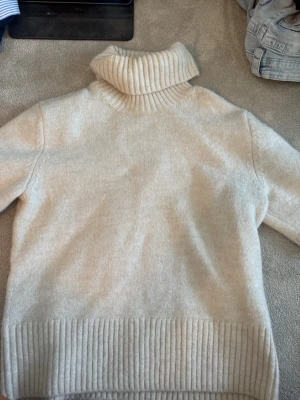 Beige polotröja i stickat material - Mysig beige stickad polotröja med bred ribbad krage, muddar och nederkant. Tröjan har lång ärm och är tillverkad i ett mjukt material som passar perfekt för kyligare dagar. Enkel och stilren design som är lätt att matcha.