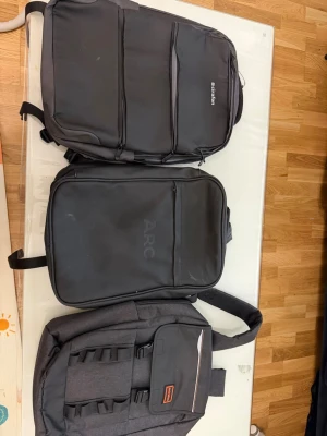 Tre svarta laptopryggsäckar - Säljer tre stilrena svarta ryggsäckar perfekta för laptop och skola. Alla har flera fack och smarta fickor för organisering. En har ARC-logga och en annan har en etikett med orange detalj. Tillverkade i slitstarkt syntetmaterial och har vadderade axelband för extra komfort.