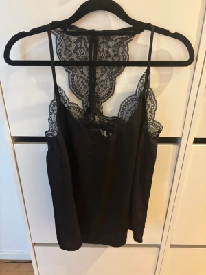 Svart linnetopp med spetsdetaljer - Säljer en svart linnetopp med tunna axelband och vackra spetsdetaljer längs urringningen och ryggen. Toppen har en elegant och feminin design med spets som ger en extra touch. Perfekt att styla till olika outfits.