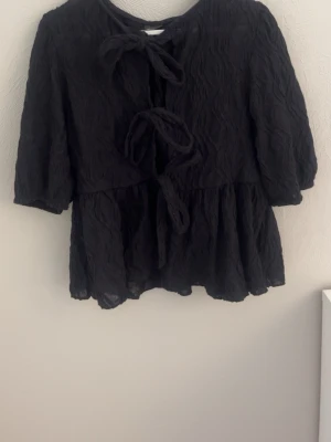 Svart blus  - Säljer en svart blus med tre knyt framtill och vågig struktur i tyget. Blusen har trekvartsärm och en lös, flowy passform som ger en avslappnad men ändå chic look. Perfekt att styla med jeans eller kjol för en trendig vibe.
