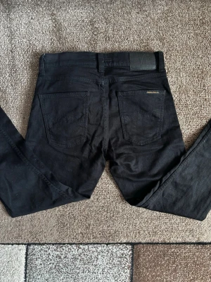Svarta jeans från Nudie Jeans - Säljer ett par svarta jeans från Nudie Jeans med klassisk femficksmodell och raka ben. Jeansen har diskreta sömmar och en patch bak i midjan. Tillverkade i slitstarkt bomullstyg och passar perfekt till en avslappnad stil. Modellen Lean dean  i storlek 30/30