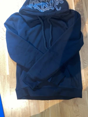 Mörkblå hoodie med tryck på huvan - Snygg mörkblå hoodie med stor huva och grova dragsnören. Huvan har ett coolt vitt tryck i graffiti-stil. Hoodien har ribbade muddar vid ärmar och nederkant. Perfekt för en avslappnad streetwear-look.