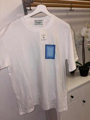 Vit t-shirt från Casablanca med blå print - Vit t-shirt från Casablanca med blått grafiskt tryck både fram och bak. Klassisk passform och rund halsringning. Tillverkad i mjuk bomull som känns skön mot huden. Perfekt för dig som gillar stilrena men ändå unika plagg.