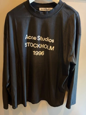 Svart långärmad Acne Studios - Svart långärmad topp från Acne Studios med vit text 'Acne Studios STOCKHOLM 1996' på bröstet. Klassisk rund halsringning och relaxed fit. Tillverkad i mjuk bomull som känns skön mot huden. Perfekt för en clean och stilren look.