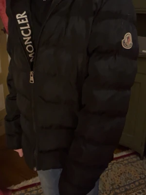 Svart pufferjacka från Moncler - Svart pufferjacka från Moncler med klassisk logga på ärmen och dragkedja framtill. Jackan har en quiltad design och är tillverkad i polyester, perfekt för kalla dagar. Snygg och stilren look som funkar till jeans och sneakers.