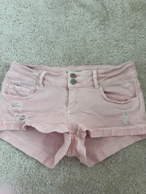 Ljusrosa denimshorts med slitningar - Säljer ett par ljusrosa denimshorts med råa kanter och slitna detaljer framtill och baktill. Shortsen har fem fickor, bälteshällor och stängs med dubbla knappar och dragkedja. Perfekta för varma dagar och ger en avslappnad vibe.💓