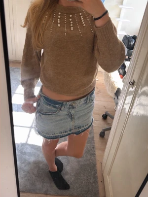Beige stickad tröja med pärlor - Supersnygg beige stickad tröja med pärldetaljer runt halsringningen. Tröjan har en croppad passform, långa ärmar och ribbade muddar. Perfekt att styla med jeans eller kjol för en trendig look.