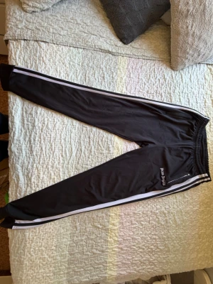Svarta mjukisbyxor Palm Angels - Säljer ett par svarta mjukisbyxor från Palm Angels med vita sidoränder och broderad logga på vänster ben. Byxorna har resår i midjan och smal passform. Perfekta för en avslappnad och trendig look.