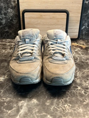 Nike Air Max 90 grå/blå sneakers - Nike Air Max 90 sneakers i grått med blå swoosh. Ovandel i mesh och mocka, klassisk Air Max-sula med synlig luftbubbla. Lågt skaft och snörning framtill. Perfekta för dig som gillar retrostil och streetwear.