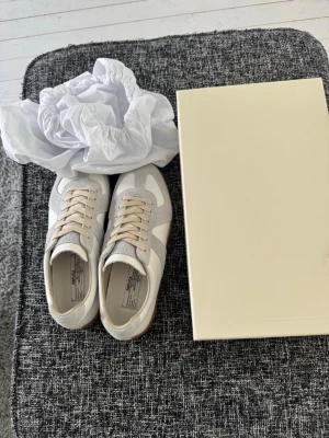 Maison Margiela vita sneakers - Tjena! Säljer ett par Maison Margiela sneakers i vitt och ljusgrått med beige snören. Skorna har en klassisk låg siluett och är tillverkade i en mix av mocka och läder. Snygg retrostil med gummisula och diskreta detaljer. Perfekta för dig som gillar minimalistisk och clean design. Kvitto finns som bevis på köp hör av er vid frågor o prisförslag ❗️🙂