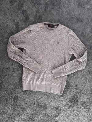 Beige kabelstickad tröja Morris - Snygg beige kabelstickad tröja från Morris med rund halsringning och diskret logga broderad på bröstet. Tillverkad i mjukt material som ger en klassisk och stilren look. Perfekt för dig som gillar tidlös design och vill ha något bekvämt. Storlek M men liten i storleken. Jag bär storlek S och den passar mig bra.