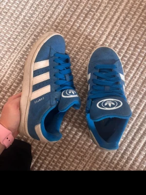 Adidas campus  - Snygga blå sneakers med vita ränder på sidorna och vit sula. Skorna har blå skosnören och en logga på plösen. Ovandelen är i mockaliknande material och sulan är i gummi. Perfekta för dig som gillar klassisk streetstil.