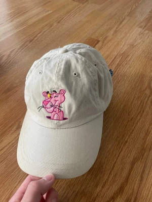 Beige keps med Rosa Pantern-brodyr - Säljer en beige keps med broderad Rosa Pantern på framsidan. Kepsen har böjd skärm, justerbar rem bak och är tillverkad i bomullstyg. Perfekt för dig som gillar tecknade klassiker och vill ha en unik accessoar. Lägg bud