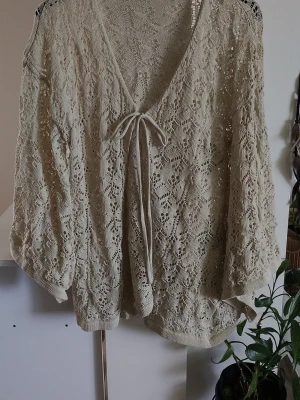 Beige virkad kofta med knytband - Supersöt beige kofta i virkad stil med vackert spetsmönster och knytband framtill. Koftan har vida ärmar och djup V-ringning, perfekt att slänga över ett linne eller klänning. Materialet känns lätt och luftigt.
