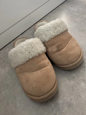 Beige tofflor med fuskpälsfoder - Mysiga beige tofflor med fluffigt vitt fuskpälsfoder. Tofflorna har en klassisk slip-in modell och ovandel i mockaimitation. Perfekta för att hålla fötterna varma hemma under kyliga dagar.