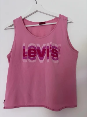 Rosa linne från Levi's - Snyggt rosa linne från Levi's med tryckt logga i vitt och olika nyanser av rosa på bröstet. Modellen är ärmlös med rund halsringning och tillverkad i mjukt bomullstyg. Perfekt för en avslappnad och trendig look.