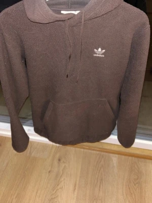 Brun teddyhoodie från Adidas - Mysig brun hoodie från Adidas i teddy-material med huva och dragsnören. Klassisk känguruficka framtill och Adidas logga broderad på bröstet. Perfekt för dig som gillar en avslappnad och trendig stil.