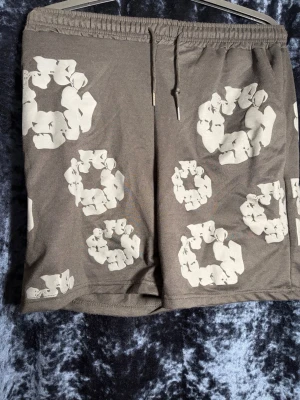 Beige shorts med blommigt mönster - Säljer ett par beige shorts med elastisk midja och snörning. Shortsen har ett unikt blommigt mönster i vitt över hela plagget och en bakficka. Perfekta för sommarens alla äventyr! (Passar S) Skickas snabbt inom 1-2 dagar. Djur- och rökfritt hem. 