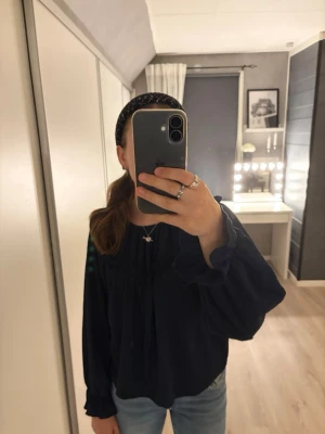 Marinblå blus  - mörkblå blus med långa ärmar. Blusen har rundad halsringning och lätt rynkade detaljer framtill. 