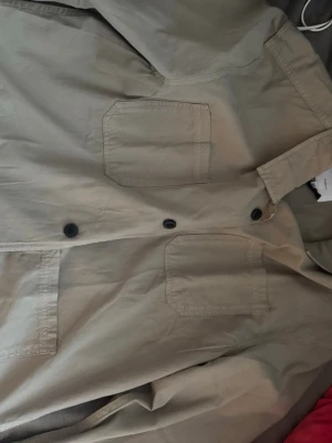 Beige overshirt från Lindbergh - Stilren beige overshirt från Lindbergh i relaxed fit. Skjortan har två stora bröstfickor med raka kanter och stängs med svarta knappar framtill. Tillverkad i ett lätt bomullstyg som passar perfekt för lager på lager.