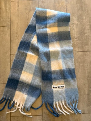 Rutig halsduk från Acne Studios - Mysig halsduk från Acne Studios i fluffig ullblandning. Den har ett rutigt mönster i blått, vitt och ljusgult samt fransar i ändarna. Perfekt accessoar för att lyfta din outfit med en trendig touch.