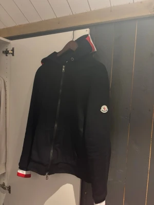 Svart Moncler hoodie  - Säljer en svart hoodie med dragkedja och huva. Tröjan har vita och röda detaljer på muddar och huva samt en patch på ärmen. Praktiska fickor framtill och ribbade muddar. Perfekt för en avslappnad och stilren look.