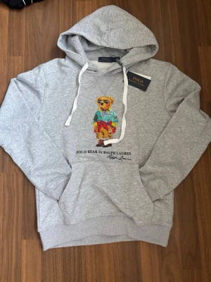Grå hoodie med Polo Bear från Ralph Lauren - Snygg grå hoodie från Ralph Lauren med Polo Bear-tryck på bröstet. Hoodien har huva med vita snören och en stor magficka. Tillverkad i mjukt bomullsmaterial och har ribbade muddar vid ärmar och nederkant. Perfekt för en avslappnad och trendig look.