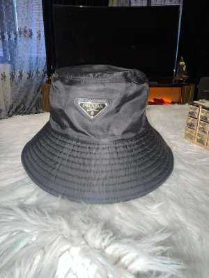 Svart bucket hat från Prada - Snygg svart bucket hat från Prada med det klassiska trekantiga metallmärket framtill. Hatten är tillverkad i ett glansigt nylonmaterial som ger en modern och trendig look. Perfekt accessoar för dig som vill sticka ut och addera lite lyx till din outfit.