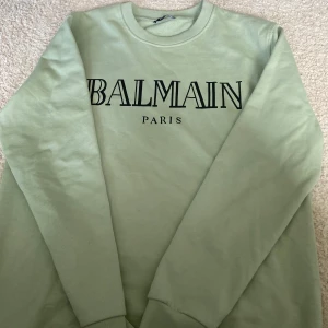 Balmain tröja - Snygg ljusgrön sweatshirt från Balmain Paris med stor svart logga framtill. Tröjan har rund halsringning och långa ärmar, perfekt för dig som gillar stilrena och trendiga plagg. Tillverkad i mjuk bomull för maximal komfort.