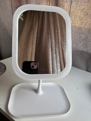 Vit sminkspegel med stativ - Stilren sminkspegel i vit plast med rektangulär form och rundade hörn. Spegeln står på ett stabilt stativ med fyrkantig fot, perfekt för sminkbordet. Enkel och modern design som passar in i de flesta rum.