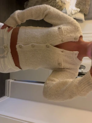 Krämvit fluffig cropped kofta - Supermjuk och fluffig krämvit kofta med cropped passform och rund hals. Koftan har dekorativa fickor framtill och stängs med pärlemorskimrande knappar. Långa ärmar och ribbade muddar ger en mysig känsla. Perfekt för lager på lager och trendiga outfits.