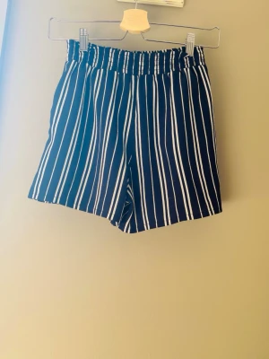 Blåa shorts med vita ränder - Snygga blåa shorts med vita vertikala ränder och elastisk midja. Perfekta för varma dagar och ger en avslappnad men trendig look. Shortsen har en loose passform och är lätta att matcha med olika toppar.
