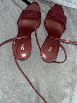 Vinröda lackpumps med remmar från Zara - Snygga vinröda pumps från Zara i glansigt lackmaterial. Skorna har spetsig tå, smala remmar som lindas runt ankeln och en elegant smal klack i metall. Perfekta för dig som vill sticka ut med en trendig och stilren look.
