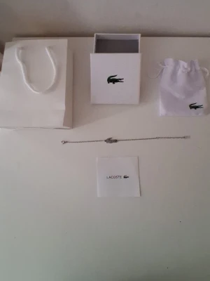 Silverfärgat armband från Lacoste - Snyggt och stilrent armband från Lacoste i silverfärg med den klassiska krokodilen som detalj. Kedjan är tunn och smidig, perfekt för dig som gillar minimalistiska accessoarer. Levereras med originalpåse och ask.