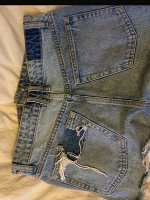 Ljusblåa jeansshorts med slitningar - Säljer ett par ljusblå jeansshorts med råa, fransiga kanter och slitningar på bakfickan. Perfekta för sommaren och ger en avslappnad vibe.