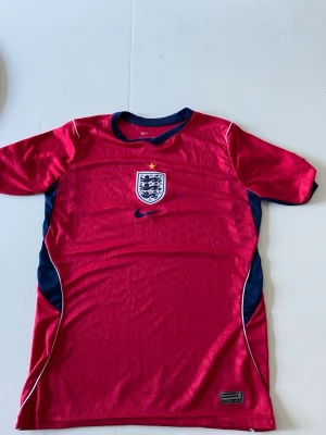 England röd fotbollströja Nike  - Säljer en röd England fotbollströja från Nike i Dri-FIT material. Tröjan har korta ärmar, blå detaljer på sidorna och runt kragen samt Englands landslagsemblem på bröstet. Perfekt för träning eller match!