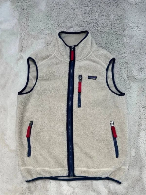 Beige fleeceväst från Patagonia med blå detaljer - Mysig beige fleeceväst från Patagonia  med marinblå dragkedjor och röda detaljer. Västen har hög krage, tre fickor med dragkedja och kontrasterande kantband. Perfekt för lager-på-lager och outdoor-vibe.