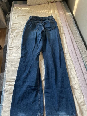 Blåa raka jeans med hög midja - Säljer ett par klassiska blå jeans med rak passform och hög midja. Jeansen har fem fickor och stängs med knapp och dragkedja. Materialet är kraftig denim i bomull och färgen är en djup blå tvätt. Perfekta till en avslappnad och trendig stil.