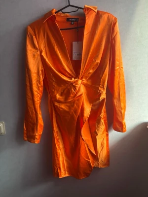 Orange satin klänning från Missguided - Snygg orange kort klänning från Missguided i glansigt satinmaterial. Klänningen har lång ärm, djup v-ringning och twistad detalj framtill. Perfekt för dig som vill sticka ut med färg och stil.