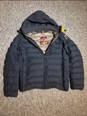 Mörkblå dunjacka från Parajumpers - Snygg mörkblå dunjacka från Parajumpers med huva och dragkedja. Jackan har en patch på ärmen, två sidofickor med dragkedja och grått foder. Perfekt för kalla vinterdagar tack vare det värmande dunet och den sportiga designen.