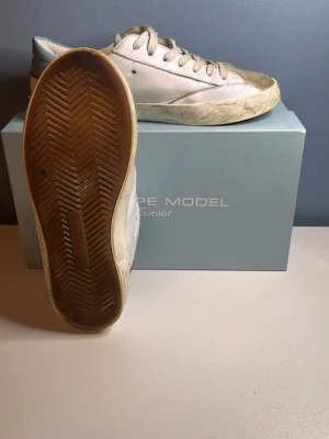 Philippe Model  KAN SÄNKA VID SNABBT KÖP - Snygga vita sneakers från Philippe Model med blå och röda detaljer på hälen. Skorna har en låg siluett, vita snören och en slitstark gummisula. Materialet är läder och designen har en cool, lite vintage look med smutsiga detaljer. 