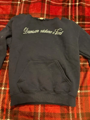 Mörkblå hoodie med text - Mörkblå hoodie med vit text 'Dansar vidare livet' på bröstet. Klassisk känguruficka framtill och ribbade muddar vid ärmslut och nederkant. Perfekt för en avslappnad stil. Materialet känns som bomullsmix. (Off shoulder)