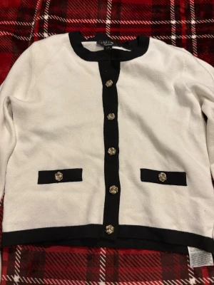 Vit och svart kofta från AMISU Knitwear - Stilren vit kofta från AMISU Knitwear med svarta detaljer längs kanter och fickor. Dekorativa guldknappar framtill och två fejkfickor. Klassisk rund halsringning och mjukt stickat material. Perfekt för en chic och enkel look. Men saknar en knapp längst upp